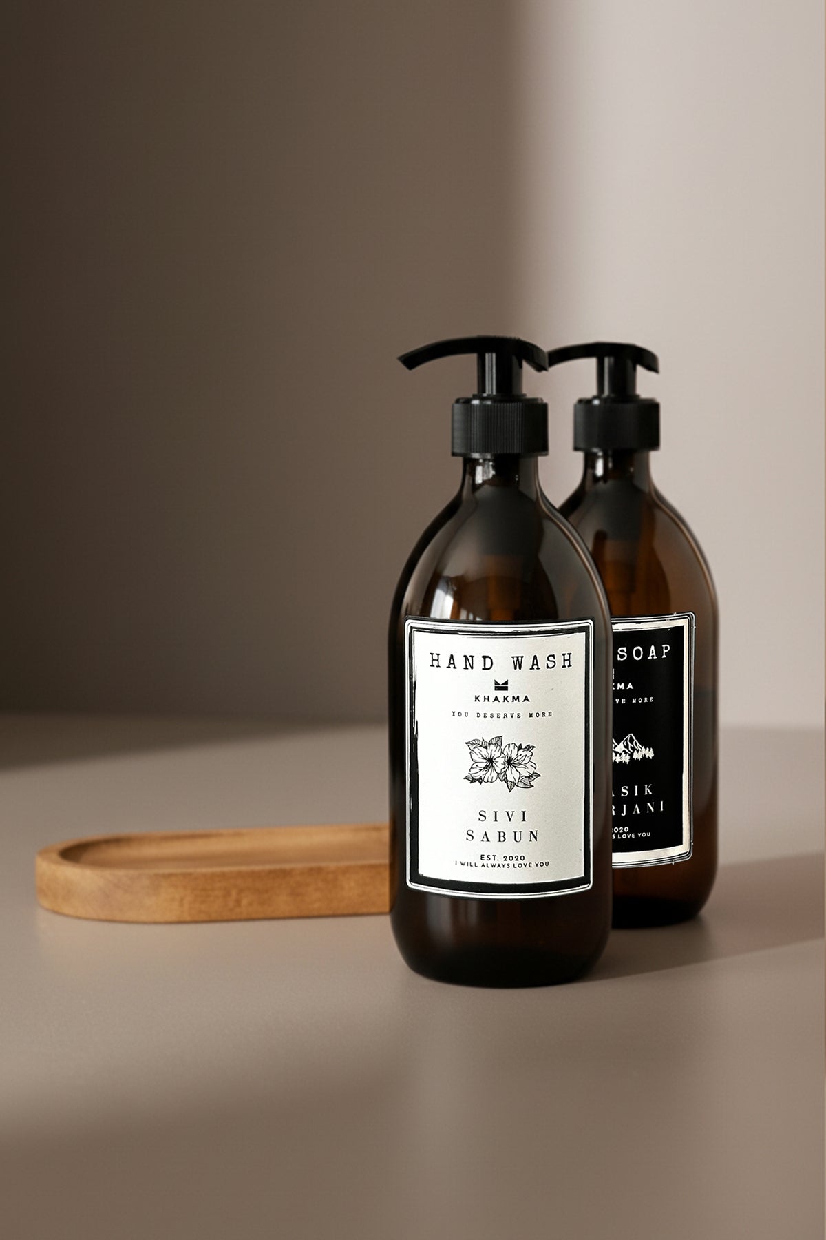 Beyaz Sıvı Sabun etiketli amber şişe önde, oval ahşap stand arkada